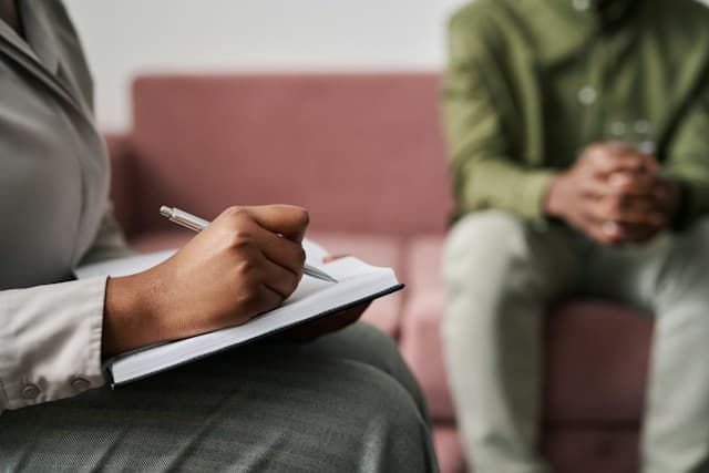 A person writing in a notebook while another person sits across from them, hands clasped.